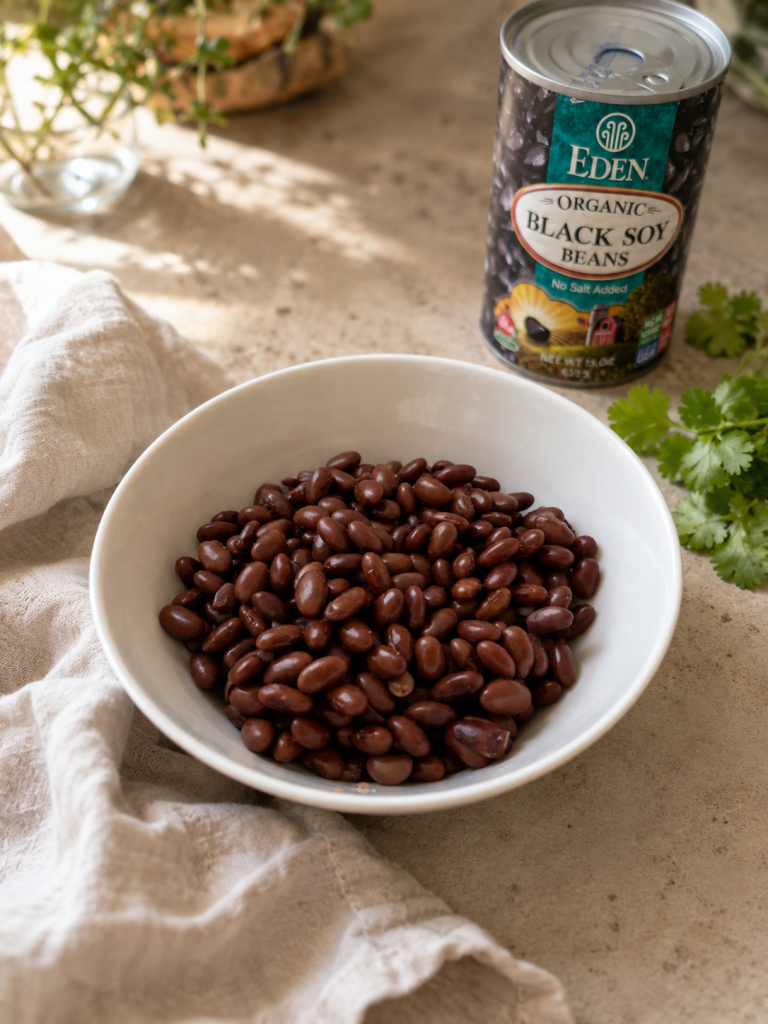Black soy beans in a white bowl with a can of Eden Organic Black Soy Beans, fresh cilantro, and a linen napkin on a light countertop.